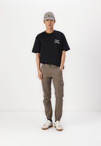 Camiseta negra de manga corta con texto blanco, pantalones cargo beiges con bolsillos laterales y zapatillas cremosas con suelas de goma, llevadas con una gorra gris.