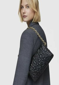 Borsa a tracolla in pelle nera con motivo impresso, tracolla in catena dorata, forma rettangolare e superficie texturizzata. Posizionata su un blazer grigio.