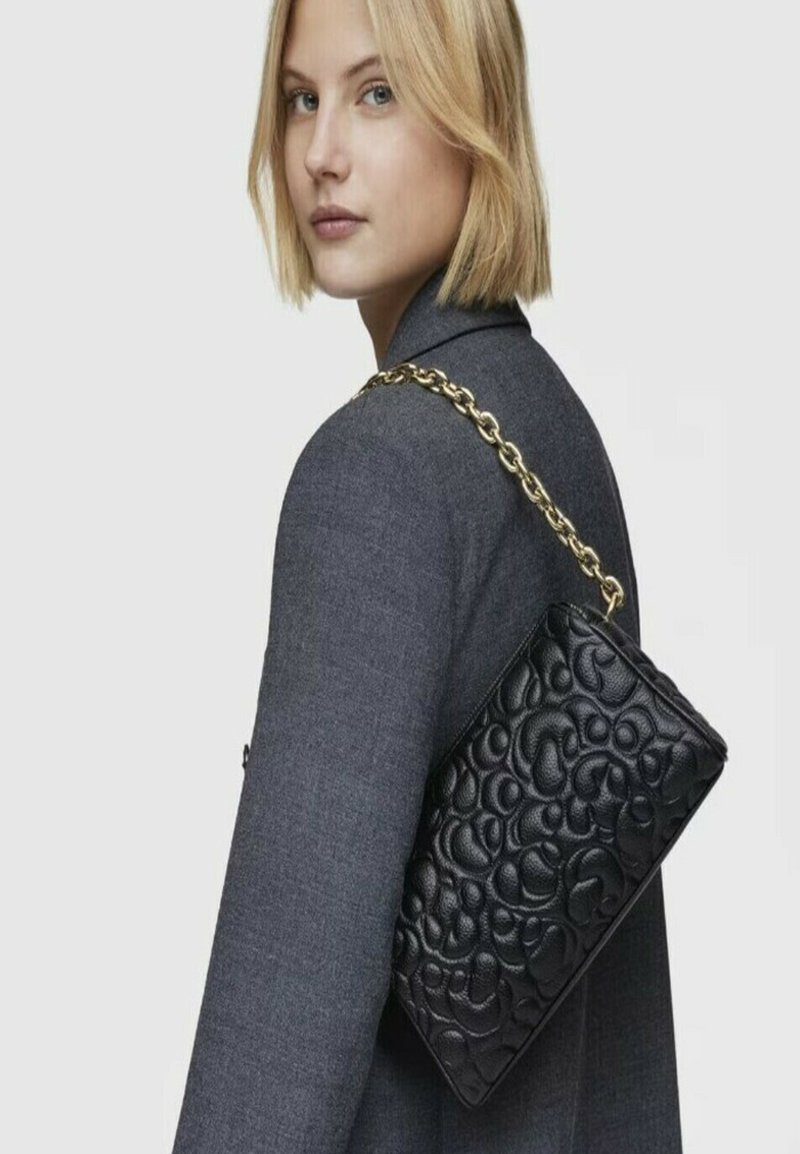 Borsa a tracolla in pelle nera con motivo impresso, tracolla in catena dorata, forma rettangolare e superficie texturizzata. Posizionata su un blazer grigio.