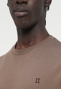 Καφέ βαμβακερό t-shirt με στρογγυλή λαιμόκοψη και κοντά μανίκια, με μικρή κεντημένη επιγραφή "II" στο στήθος. Ομαλή υφή.