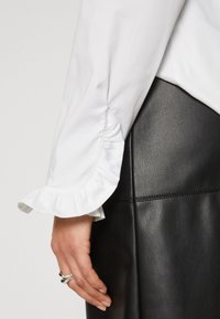 Manche de blouse blanche avec poignets froncés à côté d'une main portant des bagues en argent, associée à une jupe en cuir noire avec un détail de couture horizontale.