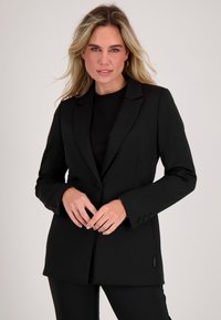 Blonde vrouw gekleed in een zwart colbert en een zwarte broek, staand tegen een effen lichte achtergrond, met handen gevouwen voor zich.