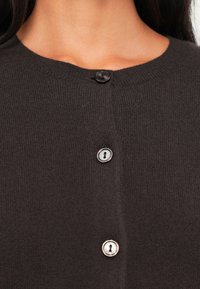 Mørkebrun cardigan med rund hals, med tre knapper: én sort og to metalliske med nøkkelhull-design. Myk strikket tekstur.