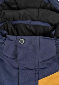 Nærbillede af en marineblå og sennepsgul jakke-krave med sort foring og en sort trykknap mærket "ISBJÖRN."