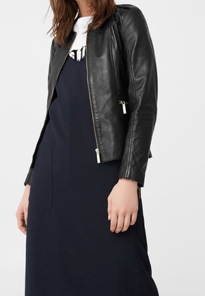 Femme portant une veste en cuir noire sur une robe bleu marine avec de grandes poches avant, debout devant un fond uni gris clair.