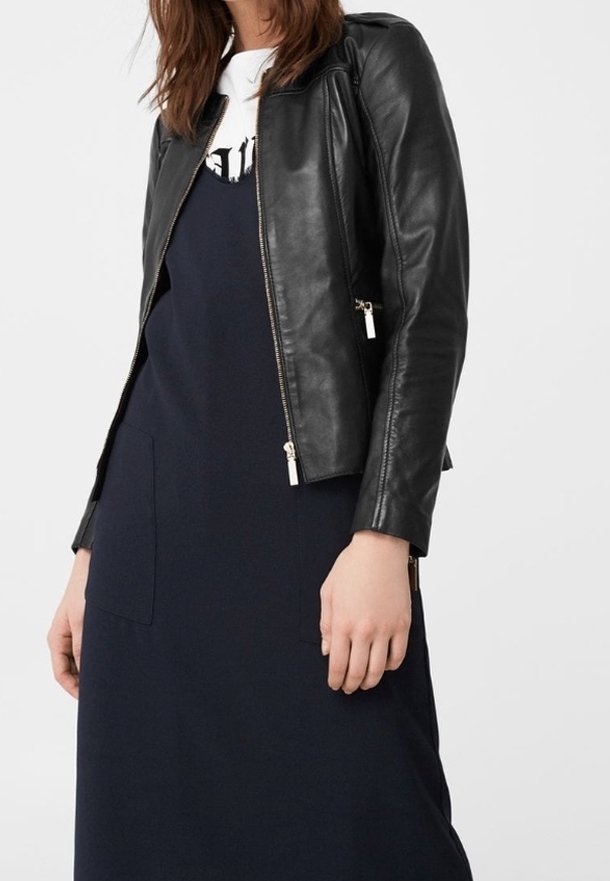 Femme portant une veste en cuir noire sur une robe bleu marine avec de grandes poches avant, debout devant un fond uni gris clair.