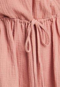 Oblačilo v prašnem roza odtenku, ki ima teksturirano, povečeno tkanino z elastičnim pasom in zavezno vezalko za prilagajanje.