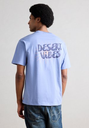 Man with afro hairstyle wearing light blue "Desert Vibes" t-shirt and blue jeans, facing away against plain background.