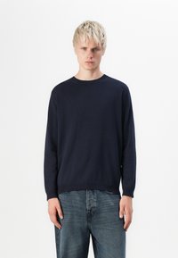 Pull bleu marine en tissu tricoté lisse, avec un col rond et des manches longues, associé à un jean bleu clair légèrement usé.