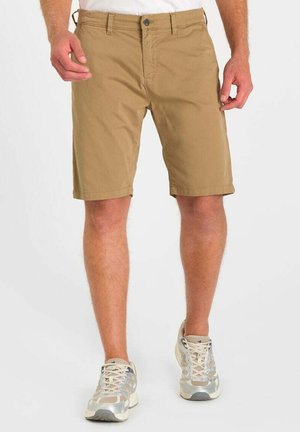 Uomo con pantaloncini beige fino al ginocchio e scarpe da ginnastica bianche-grigie in piedi davanti a uno sfondo bianco.