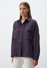 Camicia oversize in denim viola, con chiusura a bottoni sul davanti, tasche sul petto e una consistenza morbida. Abbinata a pantaloni bianchi.