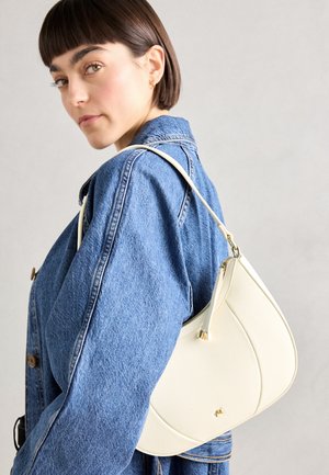 Kreminės odos petinė kuprinė su išlenkta forma ir subtiliu tekstūru, su auksine detale ant užtrauktuko, dėvima ant džinsinio švarkelio.