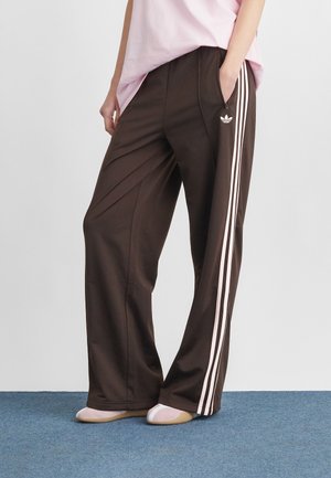 Person wearing dark brown Adidas track pants with white side stripes and logo, paired with pink top and ballet flats on blue carpet.