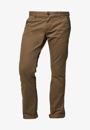 Uomo che cammina indossando una camicia bianca a maniche lunghe, pantaloni kaki, scarpe di pelle marrone e portando una tracolla di pelle marrone.