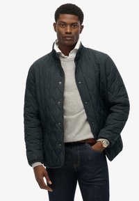 Mørk grøn kviltet jakke med høj krave, guld hardware og trykknapper, båret over en lys grå sweater og mørke jeans.