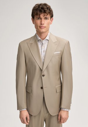 Jonge man met krullend haar, gekleed in een beige colbert over een wit overhemd met patroon, staand tegen een effen lichte achtergrond.