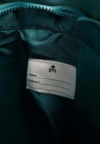 Interior de tela color verde azulado con una etiqueta blanca cosida que presenta un icono de calavera. La etiqueta tiene espacios en blanco para "NOMBRE" y "CONTACTO".