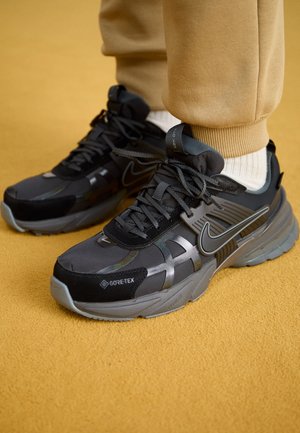 Baskets Nike Gore-Tex noires et grises portées avec un pantalon beige et des chaussettes blanches sur un tapis jaune moutarde.