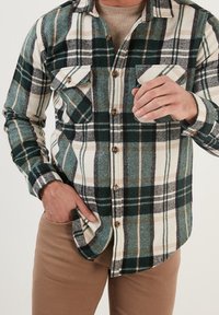 Flanellhemd mit grünem, schwarzem und cremefarbenem Karomuster, mit zwei Brusttaschen und braunen Knöpfen. Dazu beigefarbene Hose.