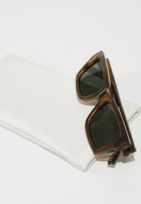 Occhiali da sole tartarugati marroni con lenti verdi, appoggiati su una custodia bianca goffrata con il logo "Le Specs". Forma angolare e finitura lucida.