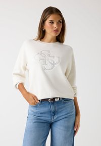 Felpa bianca con scollatura rotonda e maniche a 3/4, con una grafica del logo in paillettes argentate sul davanti. Indossata con jeans blu e una cintura marrone.