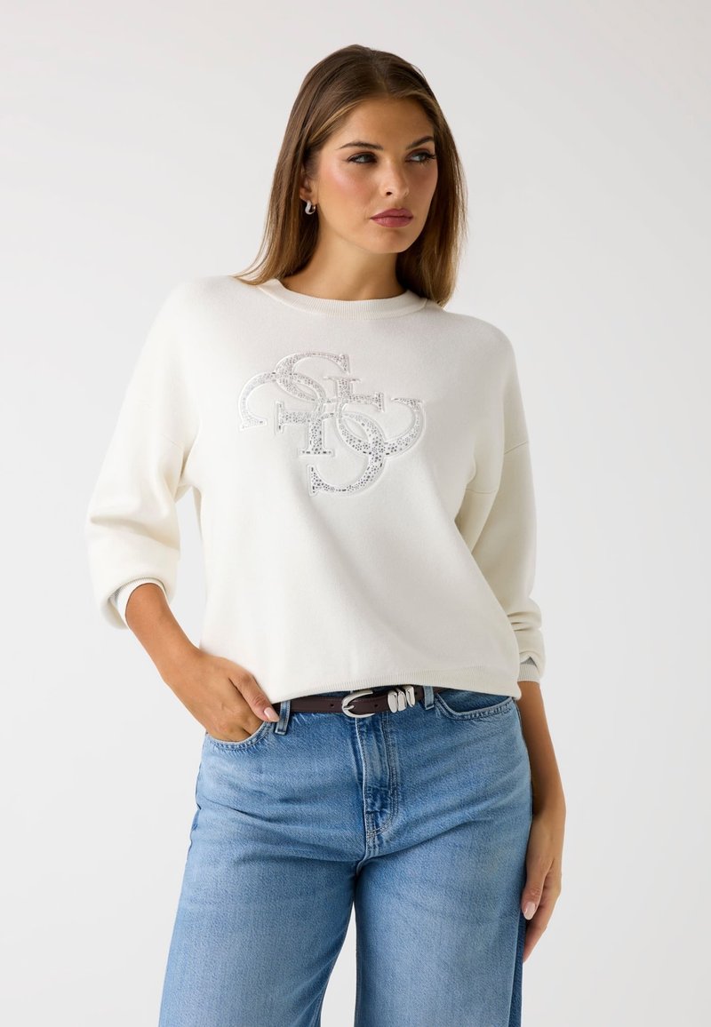 Felpa bianca con scollatura rotonda e maniche a 3/4, con una grafica del logo in paillettes argentate sul davanti. Indossata con jeans blu e una cintura marrone.