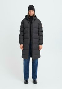 Svart pufferjakke med hette, med quiltet design og elastiske mansjetter. Båret over en svart turtleneck og blå jeans. Svart lue.