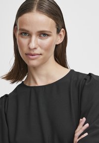 Top nero leggero con scollatura rotonda e sottili pieghe sulle spalle. Tessuto morbido, caratterizzato da un design minimalista.