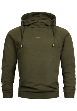 Jonge man glimlachend, draagt een olijfgroene hoodie met de tekst "ICONIC EXPERIENCE" en lichtblauwe spijkerbroek, staand tegen een effen achtergrond.
