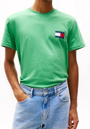 Hombre con camiseta verde de Tommy Jeans metida en unos vaqueros azul claro, con una mano en el bolsillo, contra un fondo blanco liso.
