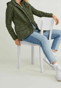 Lehká bunda olivově zelené barvy s kapucí, zapínáním na přední zip, stahováním v pase a několika kapsami, nošená přes světle modré džíny a bílé tenisky.