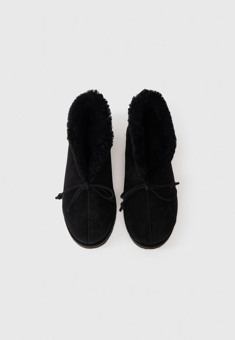 Sorte ruskinds slippers med fluffy indvendig for og bundet sløjfer, vist ovenfra på en hvid baggrund.