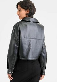 Giacca di pelle nera cropped con colletto a punta, polsini con bottoni e texture liscia. Presenta un design strutturato e dettagli minimalisti.