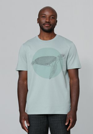 Lichtgroene t-shirt met korte mouwen, met een gedetailleerde schildpadafbeelding in grijs, geplaatst tegen een cirkelvormige muntgroene achtergrond. Zachte stoftextuur.