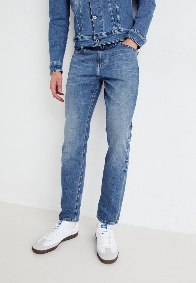Personne portant un jean en denim bleu et une veste en denim assortie, une main dans la poche, baskets blanches avec un logo bleu sur fond blanc.