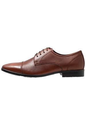 Brun læder herresko med spids tå, cap toe-design, snørelukning og lav sort hæl mod hvid baggrund.