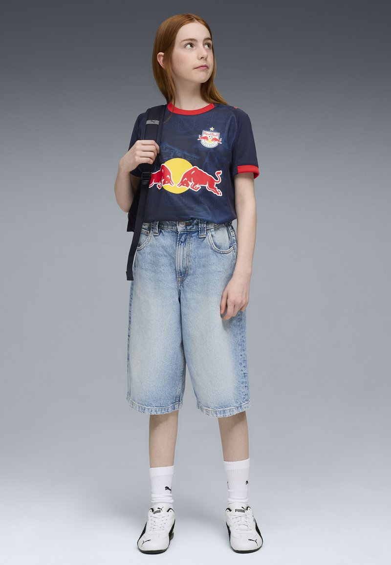 T-shirt navy avec accents rouges et logo Red Bull, associé à un short en denim léger bleu. Des chaussettes blanches et des baskets noir et blanc complètent le look.