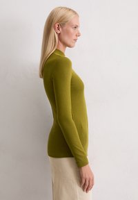 Femme aux cheveux blonds raides portant un haut à manches longues ajusté vert olive et une jupe beige côtelée, se tenant de profil contre un mur uni.