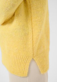 Personne portant un pull en maille jaune avec une fente sur le côté, associé à un pantalon beige clair, vue de la taille vers le bas.