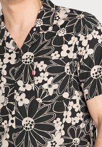 Camicia a maniche corte con bottoni nera con grandi motivi floreali bianchi. Presenta una tasca sul petto e un piccolo etichetta rossa vicino alla pattina dei bottoni.