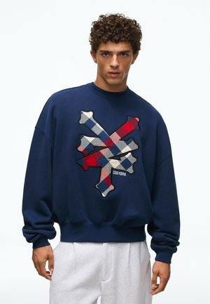 Giovane uomo con capelli ricci che indossa una felpa blu navy con una grande grafica a quadri rossi, bianchi e blu e pantaloni di colore chiaro.