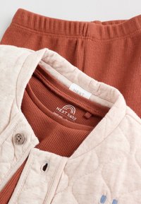 Gilet bébé matelassé beige doux sur un body bébé côtelé couleur rouille et pantalon assorti, montrant une fermeture par boutons et une étiquette de marque à l'intérieur du col.