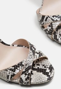 Sandales à talons hauts ouvertes avec motif peau de serpent noir et blanc et brides croisées sur fond blanc.