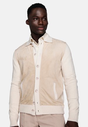 Young man wearing a beige buttoned jacket over a light shirt looking slightly to the side with a neutral background.