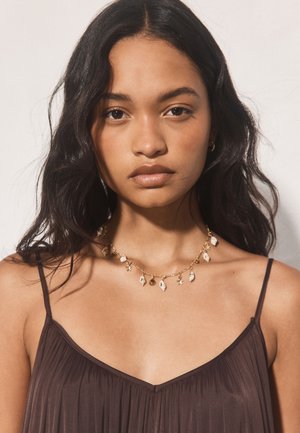 Young woman with long dark hair wearing a sleeveless brown top and a gold necklace with small seashell charms, facing forward with neutral expression.