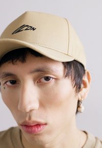 Casquette de baseball beige avec un logo noir, dotée d'un visière incurvée et de détails cousus. Le tissu semble lisse avec un design structuré.