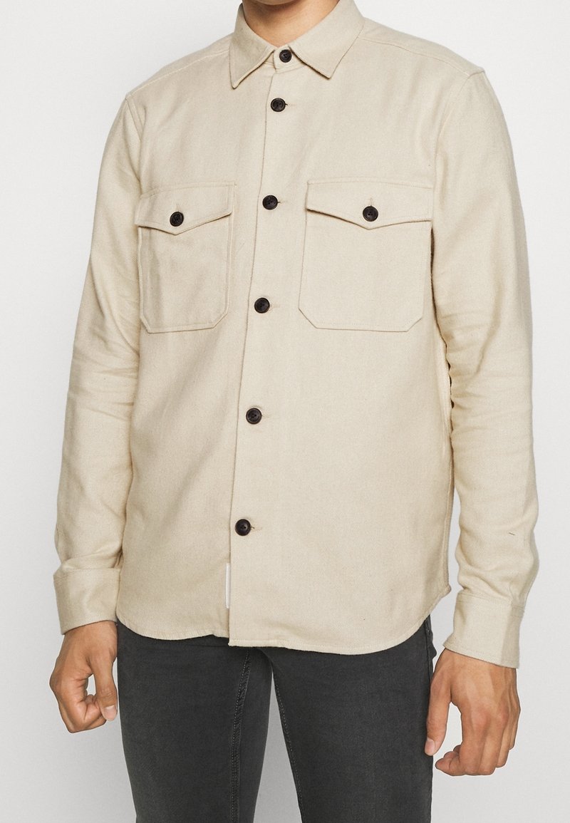 Homme portant une chemise beige boutonnée avec deux poches poitrine et des boutons noirs, associée à un pantalon gris foncé.