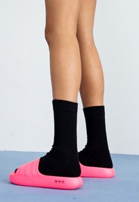 Pink slide sandals with a textured upper and open toe, paired with black ankle socks, set against a light background.