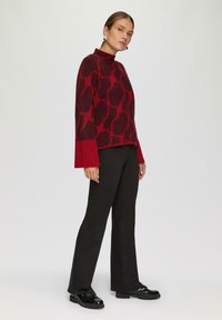 Rode pullover trui met een textuurpatroon, hoge hals en uitlopende mouwen, gecombineerd met zwarte broek en glanzende zwarte schoenen.