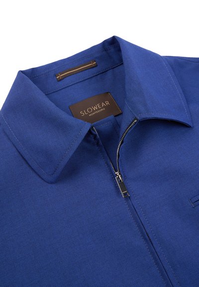 Blue zip-up shirt with pointed collar, visible silver zipper, and brown Slowear Monteodoro label inside the neckline.